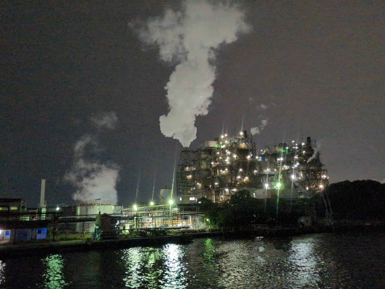 夜の運河沿いで作業灯をともす工場。大きな白い噴気の柱が空へ上がる