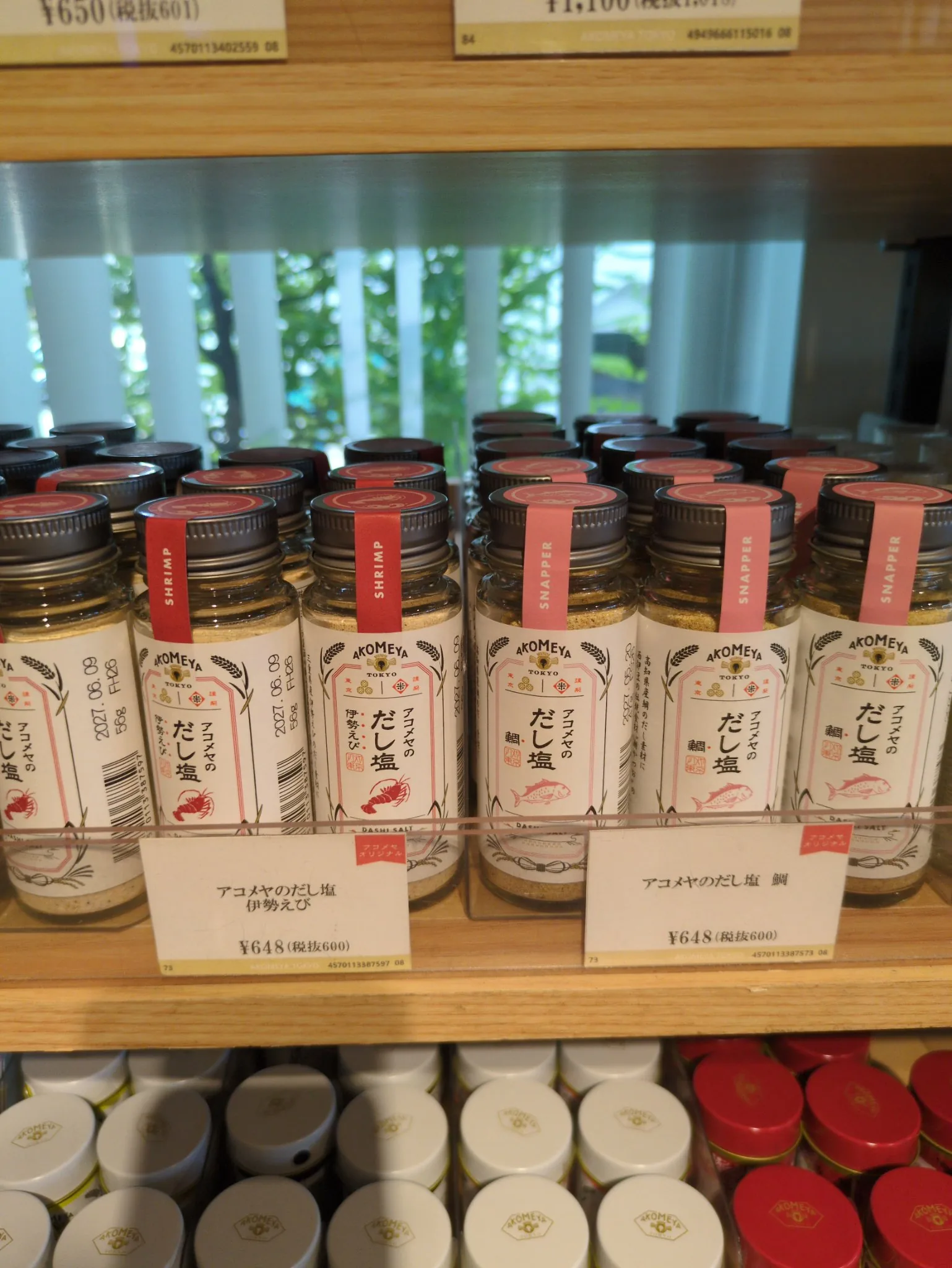 木の棚の一段に、丸い透明ガラスの瓶が左右二群に分けて、それぞれ何列も奥へ並んでいる。ラベルはどちらも同じ書式で、上にAKOMEYA TOKYOのロゴと稲穂、中央に縦書きの「アコメヤのだし塩」、その脇に赤い角印、下に赤い線画、最下部に欧文のDASHI SALT。左の群はキャップに濃い赤の帯で「SHRIMP」、線画はえび。右の群はサーモンピンクの帯で「SNAPPER」、線画は鯛。手前の値札はどちらも648円（税抜600）
