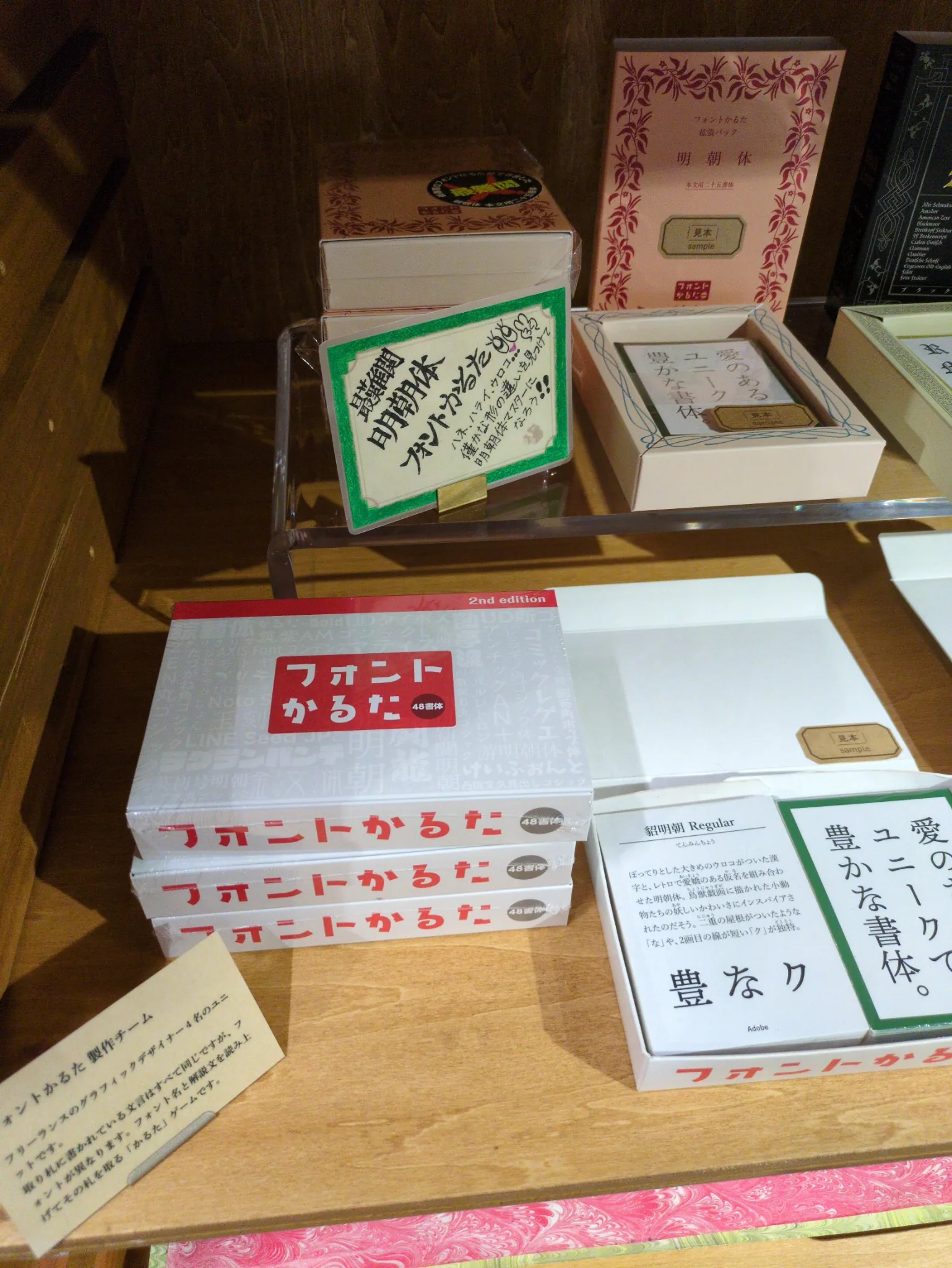 売店のフォントかるた売り場を斜め上から見たところ。手前に「フォントかるた 2nd edition 48書体」の箱が三個積まれ、右に「見本 sample」の札を添えた開封見本が置いてあり、取り札は「愛のあるユニークで豊かな書体。」が二枚と、貂明朝Regularの解説札だけが見えている。残りの取り札は箱の中にある。上段には拡張パックと、明朝体の見分け方を書いた手書きPOP