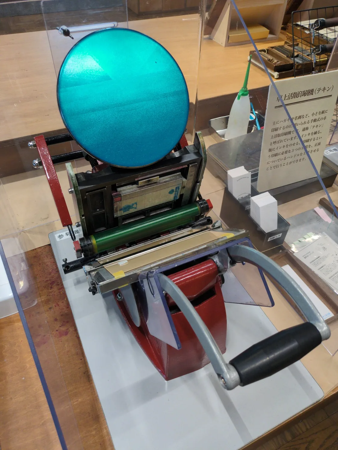 アクリルケース越しに見た卓上活版印刷機（テキン）。機体の左側に白い縦書きの札で「参号機」。いちばん上に円いインキ練り盤があり、練られているのはターコイズのインキ。その下に版を挟んだ黒い枠、中央に緑のローラー、下に赤い胴、手前に黒いグリップの大きな操作ハンドルが一本だけ突き出している。右にはこの機械の解説板と、試し刷り用の白と桃色の紙が置いてある