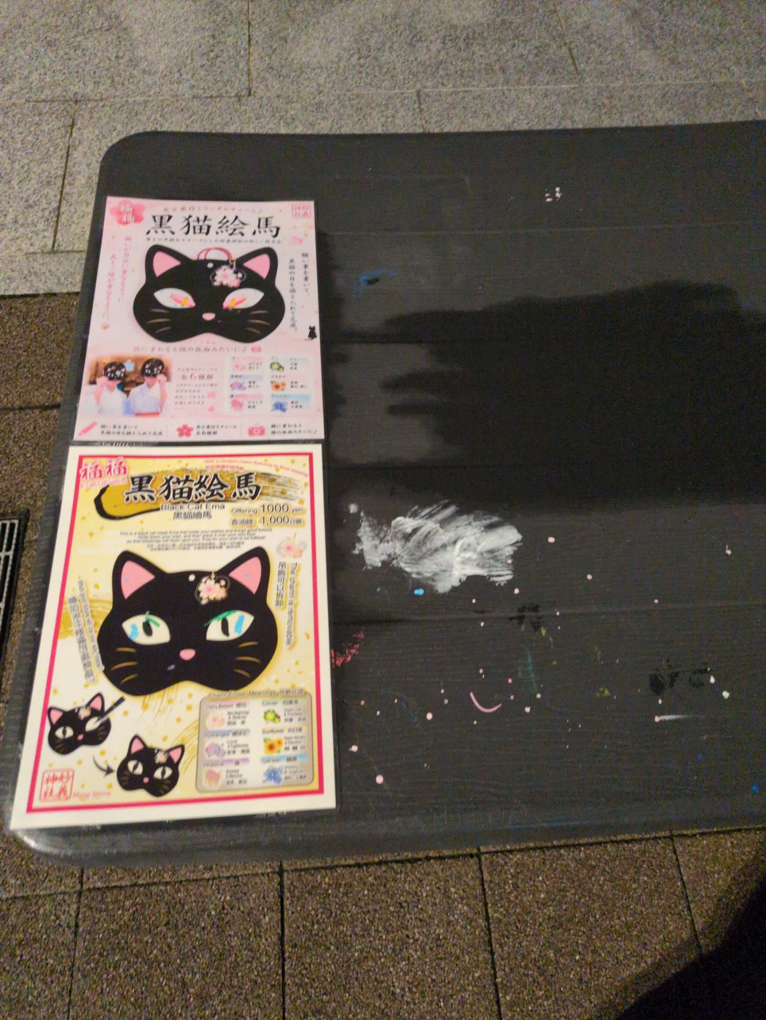 閉まった授与所の机に残るPOP。黒猫の顔型の絵馬、初穂料1,000円、全6種類のチャームの案内。机の天板には白い絵の具のこすれ跡と桃色・水色の飛沫