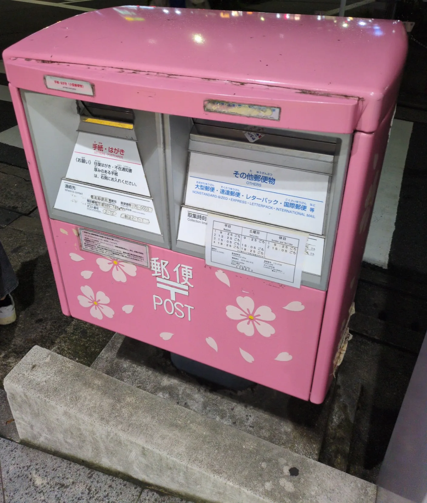 桃色の角形ポスト。白い桜の花びらが散らしてあり、「郵便 POST」の文字と2つの投函口の表示、右に取集時刻の表が読める